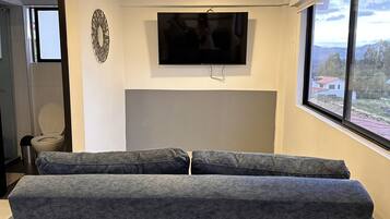 Un salon moderne avec un canapé gris, un téléviseur à écran plat fixé au mur et une grande fenêtre donnant sur l’extérieur.