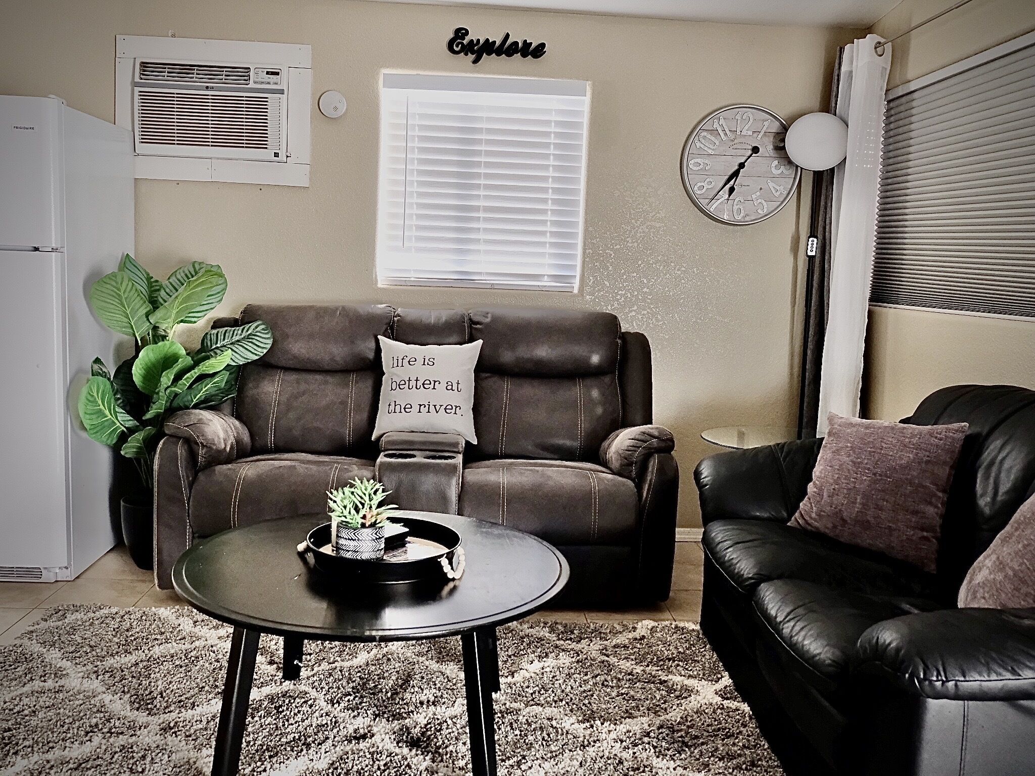 A living room with a brown leather sofa, a small round table with a tray and plants, a white air conditioner, a window with blinds, and a wall clock.