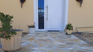 Une entrée dallée de pierres, avec une porte blanche, des plantes en pot et des paniers fixés au mur.