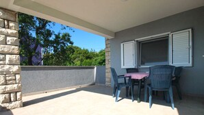 Eine überdachte Terrasse mit Tisch und Stühlen, ein Fenster mit Rollläden und eine Steinmauer.