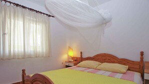 Ein Schlafzimmer mit einem hölzernen Bett, einem Nachttisch mit Lampe, einem Fenster mit durchsichtigen Vorhängen und einem Moskitonetz über dem Bett.
