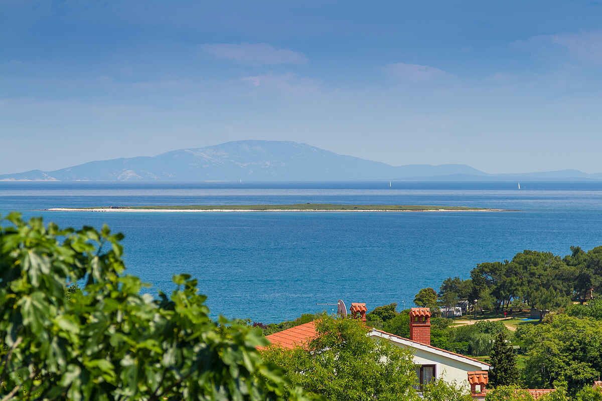 A coastal view with a house in the foreground, a clear blue sea, and a mountain in the background.