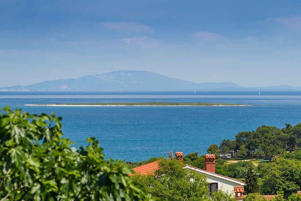 A coastal view with a house in the foreground, a clear blue sea, and a mountain in the background.