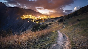 A valley with a path leading to a lit-up town nestled in the mountains.
