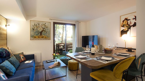 A modern living room with a blue sofa, a coffee table, and a dining area with a table set for two. There is a painting on the wall and a TV on the opposite wall.