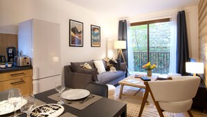 A modern living room with a sofa, coffee table, and dining area. There are framed artworks on the wall, a lamp, and a view of greenery outside.