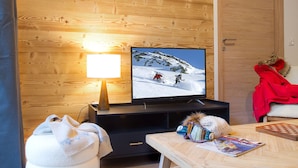 A cozy living room with a wooden wall, a flat-screen TV, a lamp, a table with a magazine, and a red jacket on a chair.
