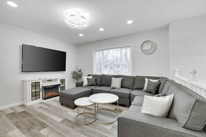 Uma sala de estar moderna com um sofá seccional, uma TV na parede, uma lareira e uma grande janela com vista para uma casa.