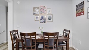Un comedor con mesa de madera y cuatro sillas, un florero con flores, y una pared con obras de arte enmarcadas y un cartel.