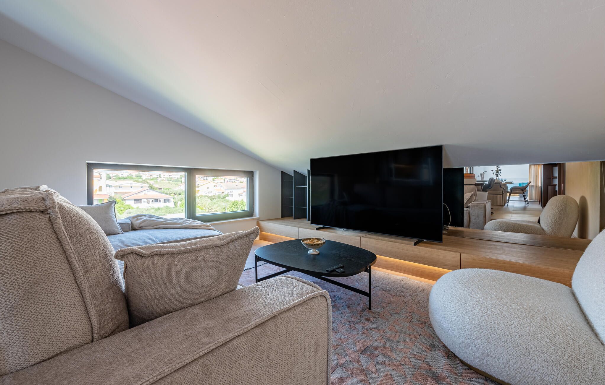 A modern living room with a large flat-screen TV, comfortable seating, and a view of the outside through large windows.