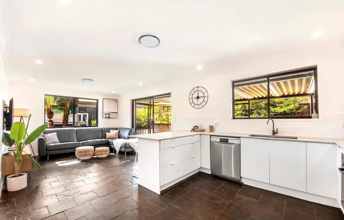 A modern kitchen with white cabinetry, a built-in dishwasher, and large windows offering a view of greenery.