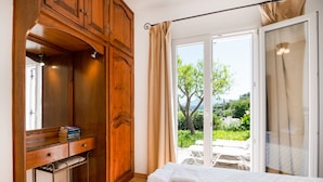 A bedroom with a wooden wardrobe, a bedside table with books, a bed with a white duvet, and a sliding glass door leading to an outdoor area. - Spiros in Saint Stephanos, Corfu - Idyllic Mediterranean Retreat (Saint Stephanos N/E)