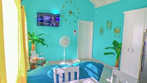 A room with a teal wall, a white door, a TV mounted on the wall, a fan, and a table with chairs. - A Blissful Carib/n Getaway! Work or Leisure, 15 mins to Beaches & Restaurants. (Saint John's)