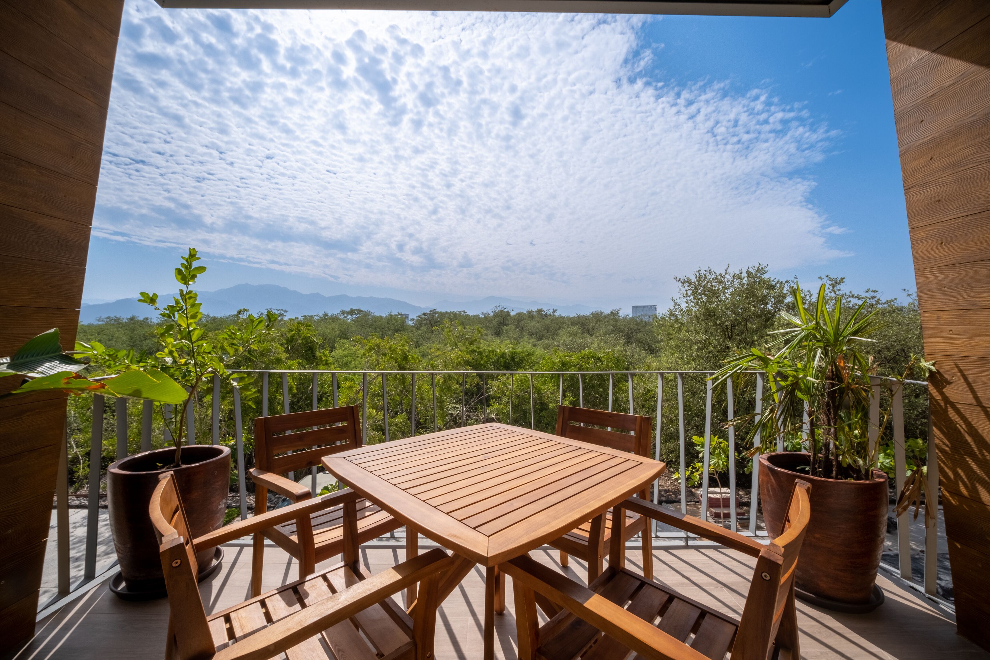 Um deck de madeira com mesa e cadeiras, rodeado por plantas em vasos, com vista para uma paisagem exuberante com árvores e montanhas.