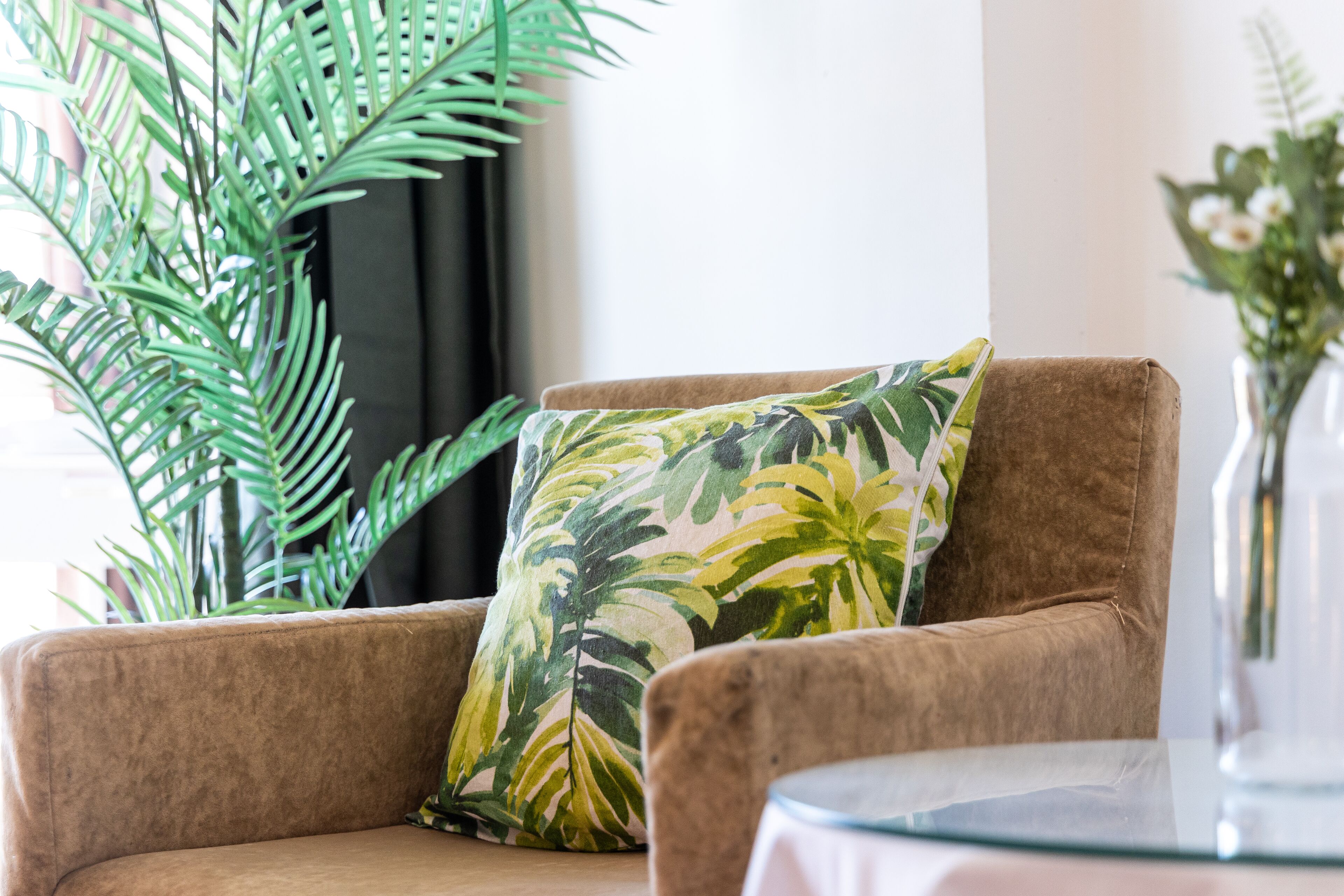 Ein braunes Sofa mit einem tropischen Blattmuster-Kissen, einem Glastisch und einer großen, grünen Pflanze im Hintergrund.