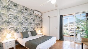 A bedroom with a bed, nightstands, a view of the city through a large window, and a balcony with a chair. - Holidays2Arroyo de la Miel Hercules (Benalmádena)