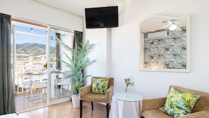 A modern living room with a sofa, armchair, and a small table. There is a large window with a view of a city and mountains. - Holidays2Arroyo de la Miel Hercules (Benalmádena)