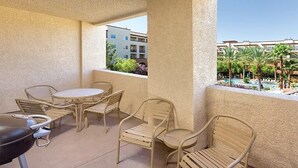 Balcón con mobiliario exterior, que incluye una mesa y sillas, con vista a un área de piscina con palmeras y un edificio al fondo.