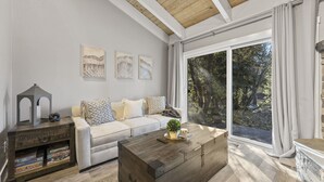 A living room with a sofa, coffee table, and a sideboard with board games. There are large windows with curtains and a view of trees outside.