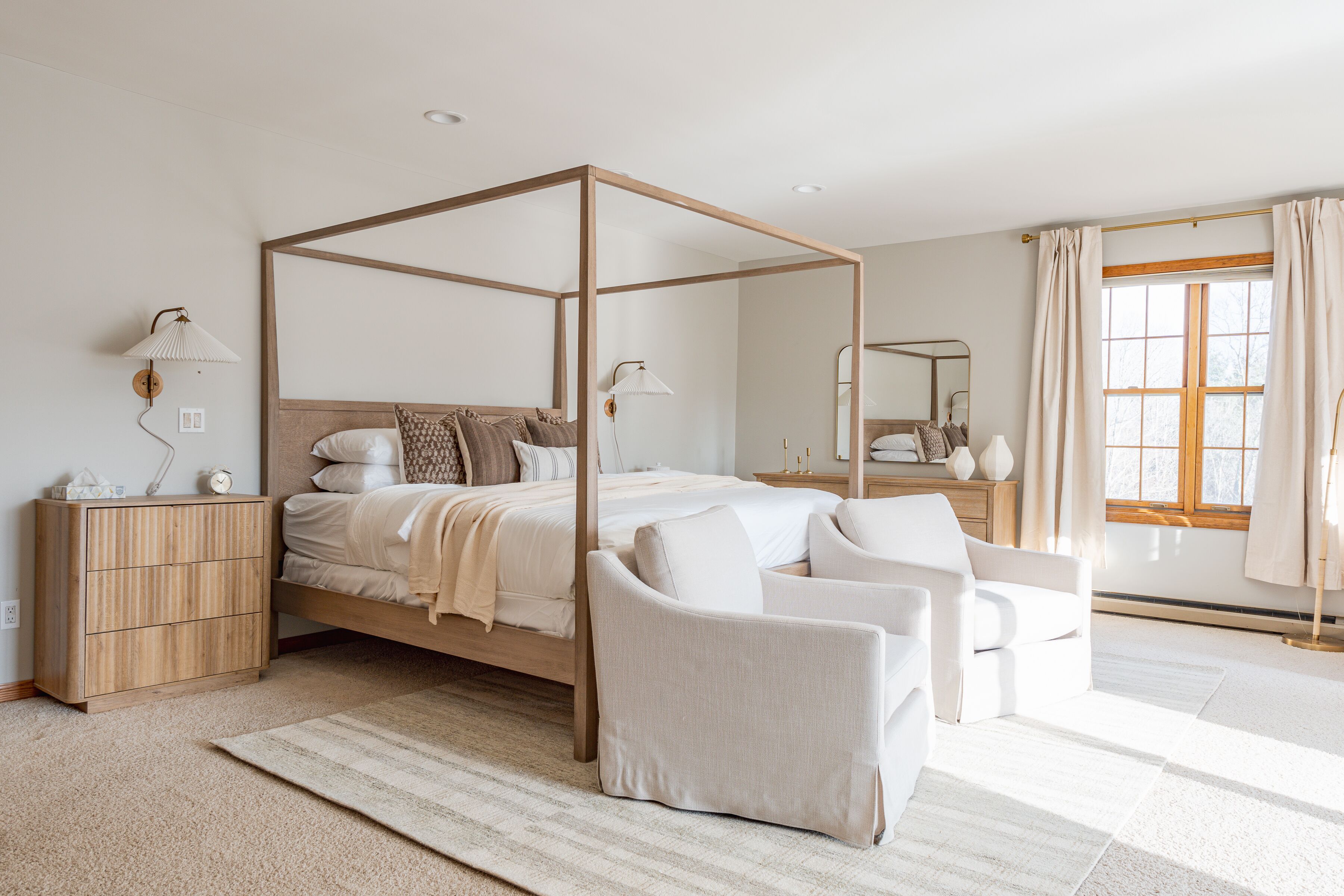 A four-poster bed with a wooden frame, a bedside table with a lamp, a chair, and a large window with curtains.
