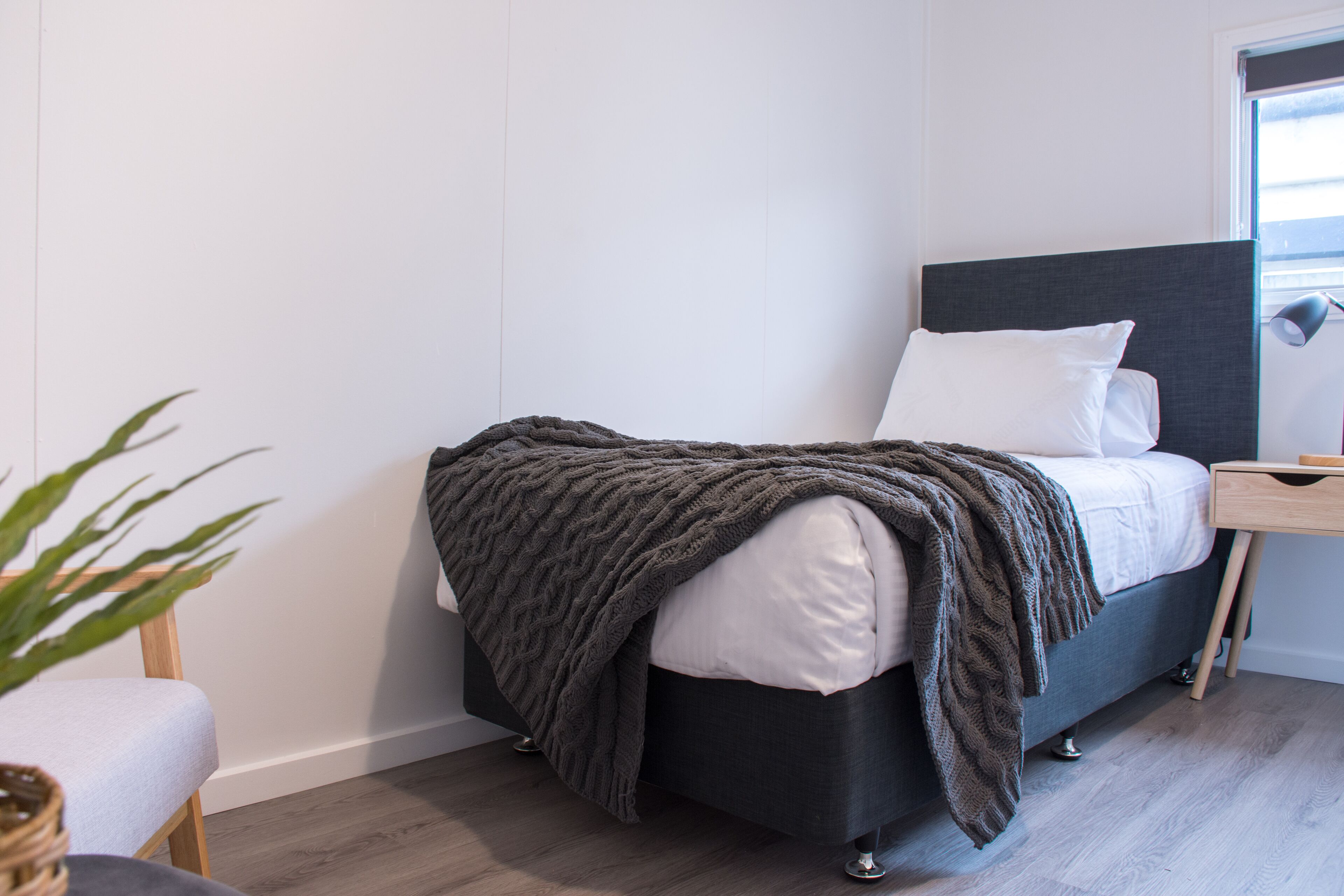 A modern bedroom with a dark headboard, a bedside table, and a window with blinds.