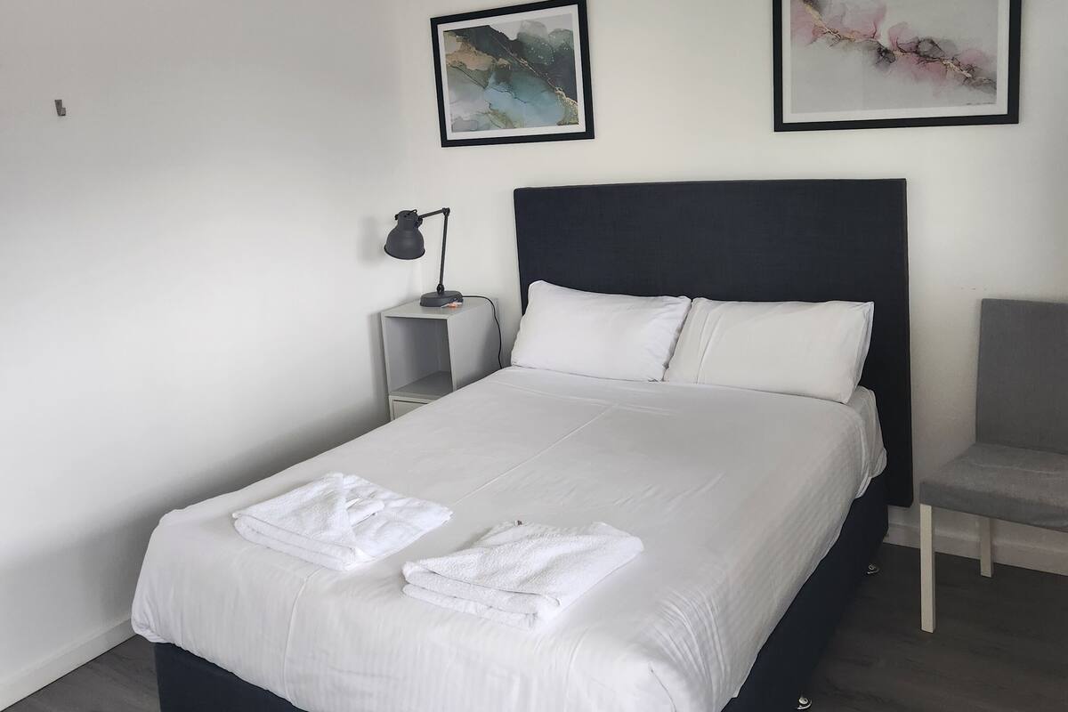 A neatly made bed with white linens and towels, a black headboard, and two framed abstract paintings on the wall.