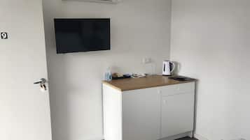 Ein kleiner, sauberer Raum mit einer weißen Tür, auf der '9' steht, einer Holzauslage mit einem Wasserkocher, einem kleinen Tisch und einem an der Wand montierten Fernseher.