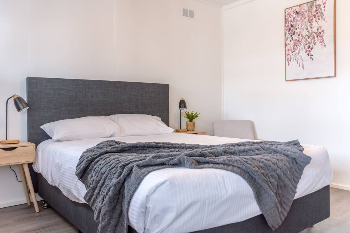 A modern bedroom with a grey headboard, white bedding, a grey knitted throw, a bedside lamp, a small plant, and a framed floral artwork on the wall.