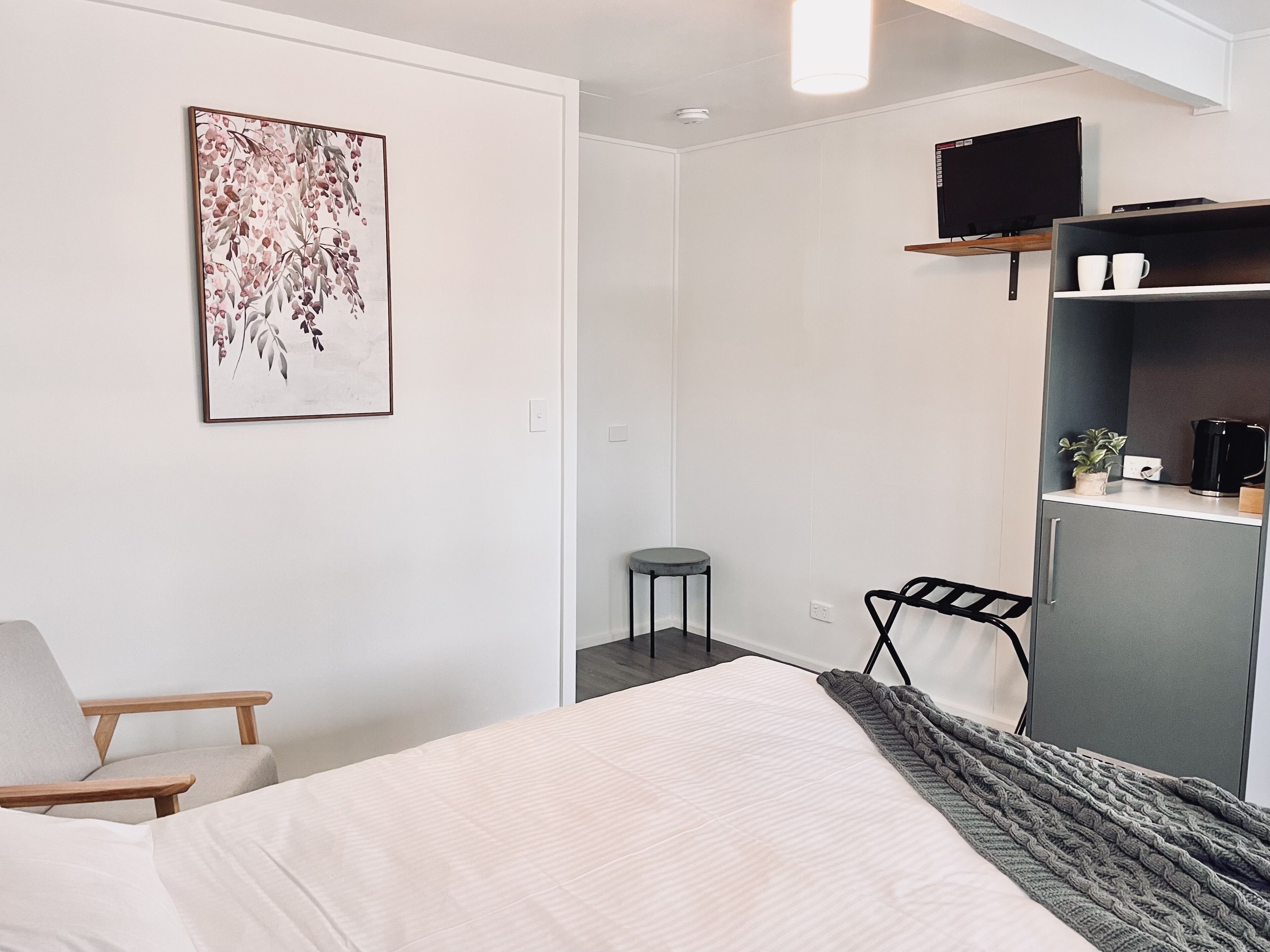 A modern hotel room with a bed, a chair, a desk, a television, and a shelf with cups and a plant.