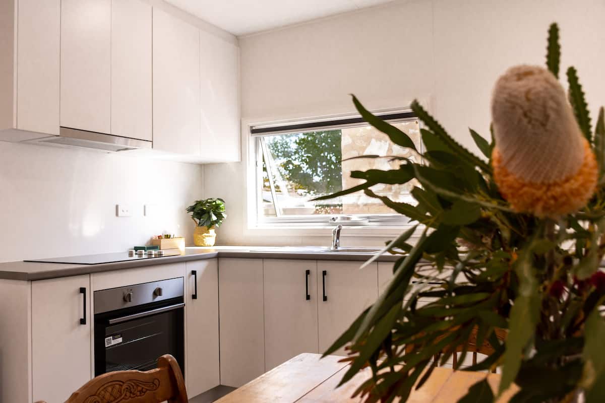 A modern kitchen with white cabinets, a wooden dining table, and a window with a view of trees.