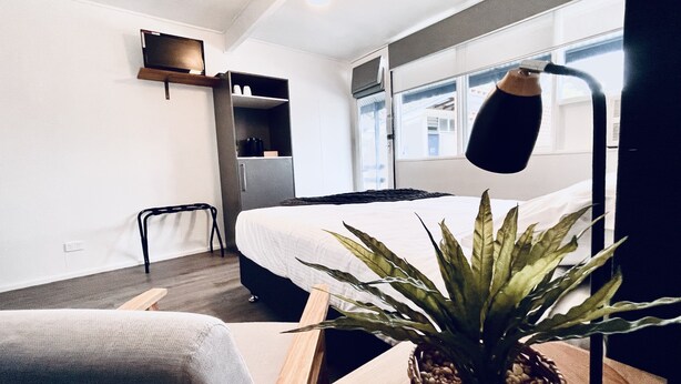 A modern hotel room with a bed, a small desk with a plant, a chair, a shelf with cups, and a window with a view of buildings. - Motel on Becker (Tintinara)