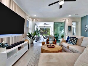 A modern living room with a flat-screen TV, a sofa, a coffee table, and a dining area with outdoor seating.