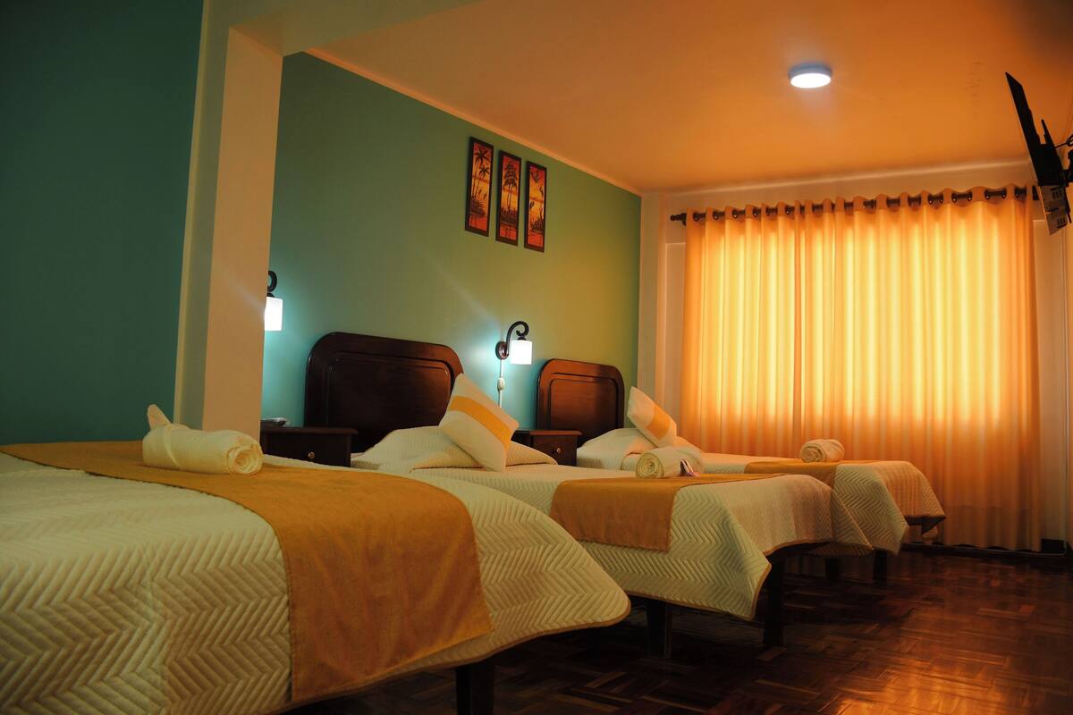 A hotel room with three beds, each with a headboard and a blanket. There are wall-mounted lights and a ceiling light.