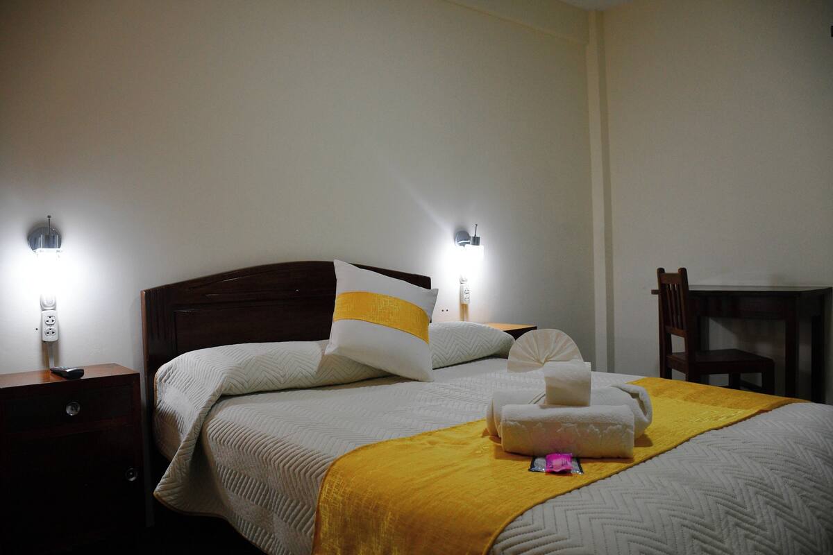 A neatly made bed with a yellow throw blanket, white pillows, and a white towel on the side. There are two wall-mounted lights on either side of the bed.
