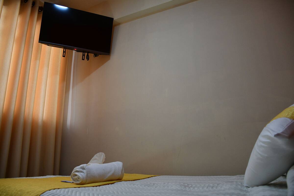 A hotel room with a bed, a flat-screen TV mounted on the wall, and a window with curtains.