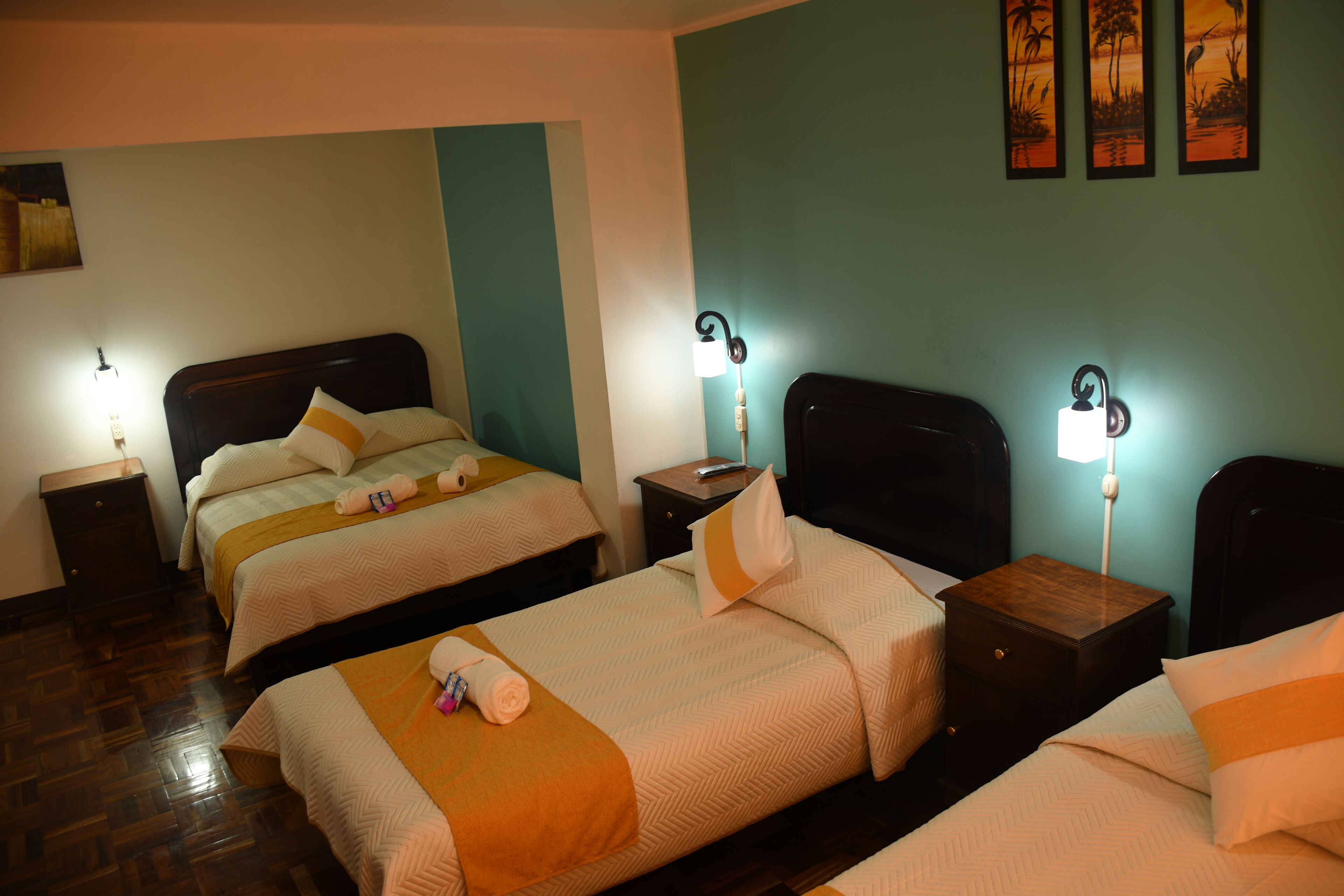 Quarto de hotel com duas camas, cabeceiras de madeira, mesas de cabeceira com abajures e uma parede de destaque em tom de verde-água.