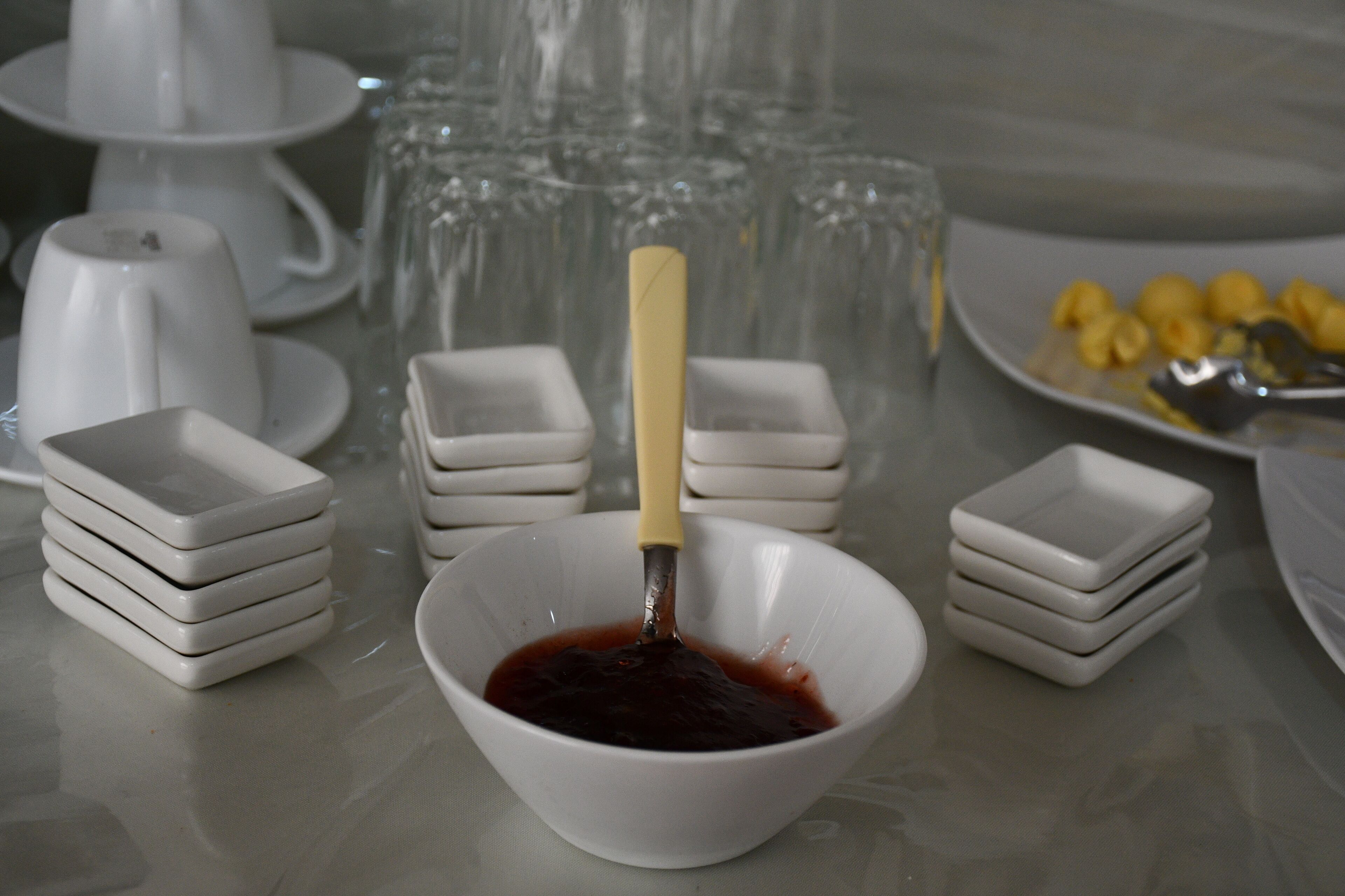 A table with a bowl of red sauce, stacked white dishes, and a plate of yellow items.
