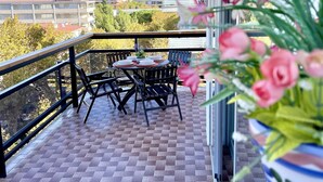 A balcony with a table and chairs, a potted plant with pink flowers, and a view of buildings and trees.