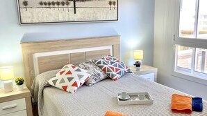 A neatly arranged bedroom with a bed, pillows, a bedside table with a lamp, a tray with cups, and a window with a view.