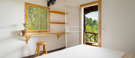 A bedroom with a large bed, a window with a view of greenery, a fan, and a wooden shelf with a plant.