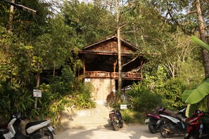 A rustic wooden structure with a porch, surrounded by dense greenery and a dirt path leading to it. - The Source Koh Phangan (Ko Pha-ngan)