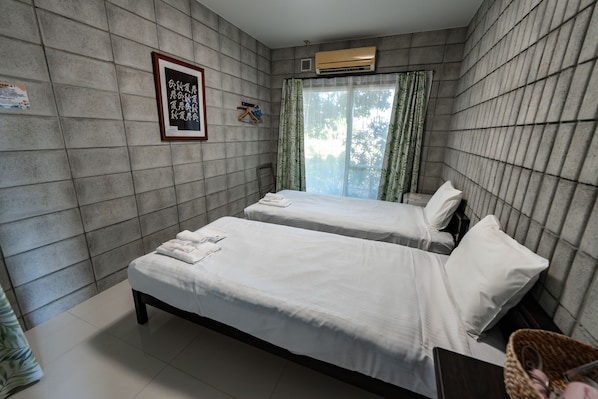 A hotel room with two single beds, a window with curtains, an air conditioning unit, and a framed picture on the wall.