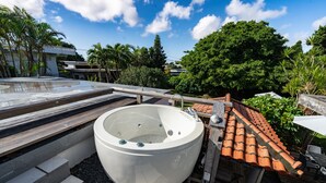 Un jacuzzi en una terraza en la azotea con escalones que llevan a él, rodeado de vegetación y un cielo despejado.