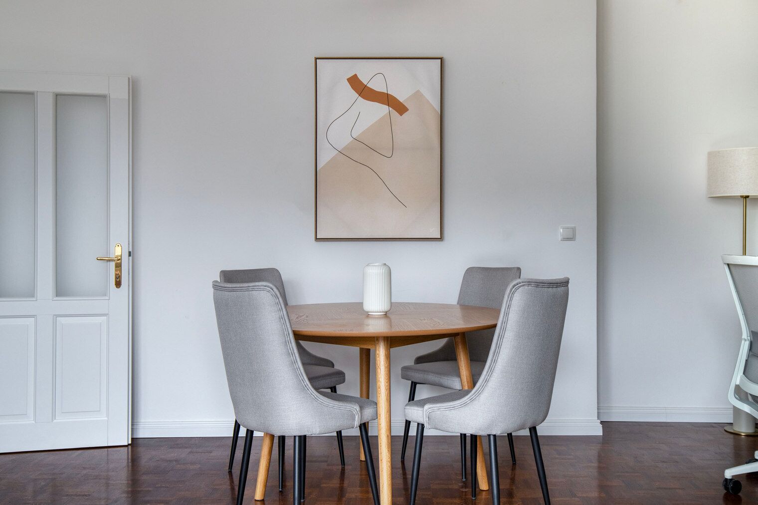 A dining area with a round table, four grey upholstered chairs, a white vase, and a framed abstract painting on the wall.