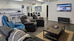 Un salon moderne avec un téléviseur à écran plat, des fauteuils inclinables, une table basse et un coin cuisine à l’arrière-plan.