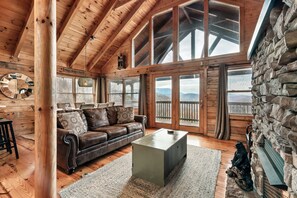 A cozy cabin interior with a wooden ceiling, large windows, a leather sofa, and a stone fireplace.