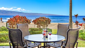 A round table with wicker chairs and a bottle of wine, overlooking a beach with a view of the ocean and a distant mountain range. - Maui Resort Rentals: Kaanapali Ali'i – Oceanfront Ground Floor + Yard Area, 6 Total Bedrooms+DEN, Sleeps 20, Steps to Pools, Beach, BBQs! (Lahaina)