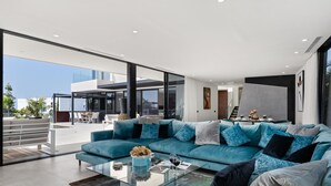 A modern living room with a large teal sectional sofa, a glass coffee table, and a view of the outdoor patio area. - KARAT Villa Serendibida (Playa Paraiso)