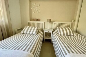Quarto duplo com duas camas, cada uma com roupa de cama listrada, uma pequena mesa de cabeceira com abajur e um objeto decorativo na parede.