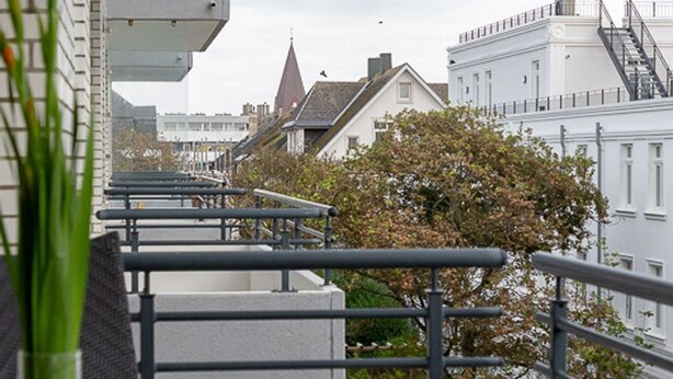 Ein Balkon mit Metallgeländern, der auf eine Reihe von Gebäuden mit traditioneller Architektur blickt, darunter ein Kirchturm.